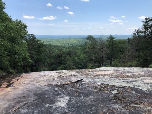 A rocky outcrop overlooks a lush green valley and distant hills under a blue sky with scattered clouds.