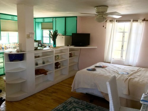 A cozy room with a bed, open shelving, a TV, and a kitchenette in the background, featuring bright colors and natural light.