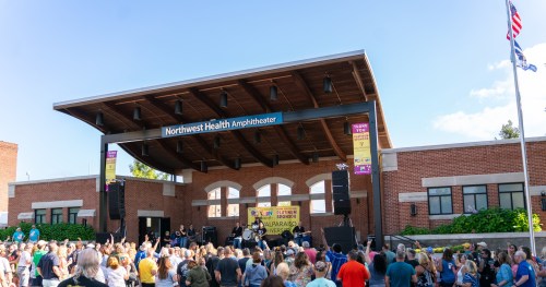 A large crowd gathers at the Northwest Health Amphitheater for a live outdoor concert under a clear blue sky.