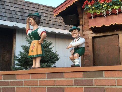 Two colorful wooden statues in traditional Bavarian attire, one woman waving and one man in a playful pose.
