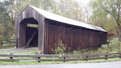 A rustic wooden covered bridge surrounded by trees and a fence, set in a peaceful rural landscape.