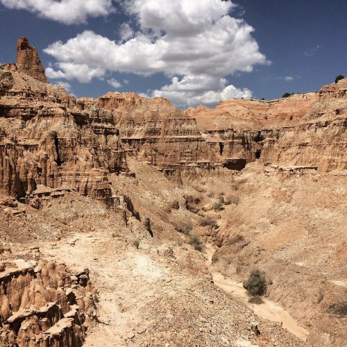 A vast, rugged canyon landscape with steep, rocky cliffs and scattered clouds in a blue sky.