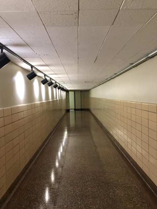 A long, empty hallway with lights on the ceiling and tiled walls, leading to a closed door at the end.