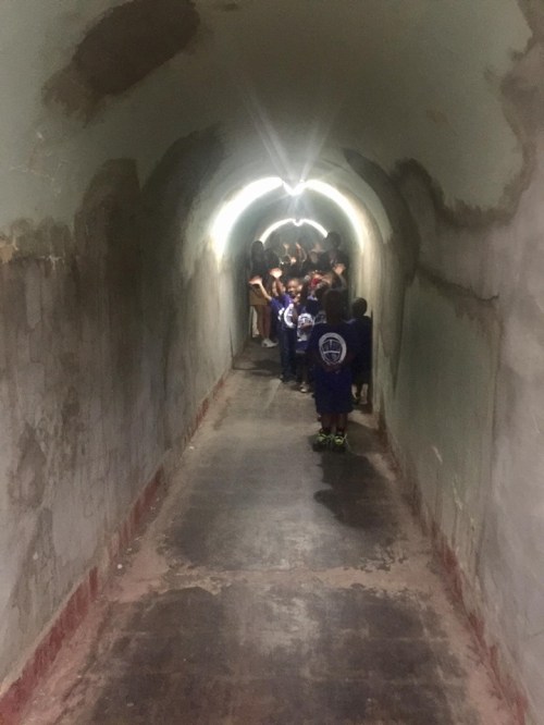 A dimly lit tunnel with a group of people walking towards the light at the end, surrounded by peeling walls.