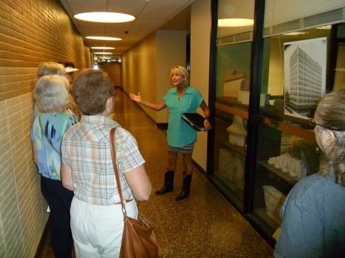 A group of women listens to a speaker in a hallway, with a building photo displayed in the background.