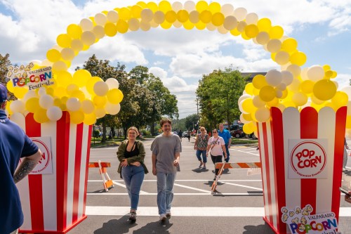 A colorful balloon arch marks the entrance to the Valparaiso Popcorn Festival, with two people walking underneath.