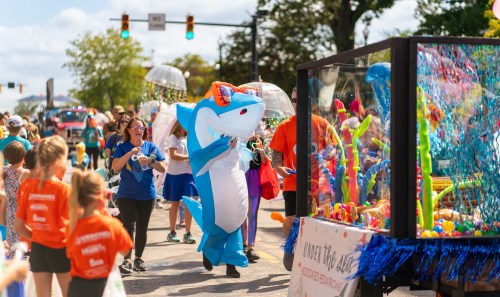 A parade scene with a person in a shark costume, colorful decorations, and people enjoying the festivities.