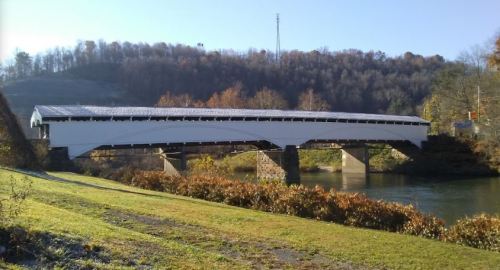 A white covered bridge spans a river, surrounded by green grass and autumn foliage under a clear blue sky.