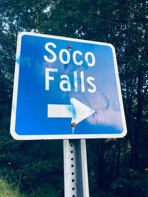 Blue sign with white text reading "Soco Falls" and an arrow pointing to the right, surrounded by greenery.