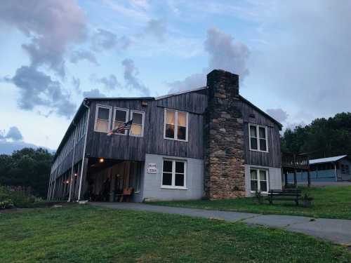 A rustic wooden building with a stone chimney, surrounded by grass and trees, under a cloudy sky.