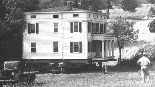 A historic house being moved on a flatbed truck, with a man walking nearby in a rural setting.