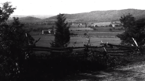 A black and white landscape featuring rolling hills, fields, and a rustic wooden fence in the foreground.