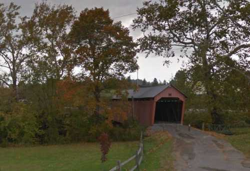 A red covered bridge surrounded by trees with autumn foliage, set against a cloudy sky.