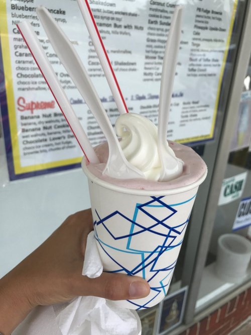 A hand holding a cup of pink soft serve ice cream topped with whipped cream, featuring multiple spoons and straws.