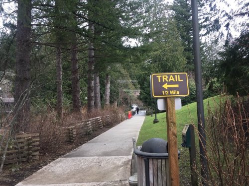 A sign points left to a trail, indicating it's half a mile, alongside a paved path surrounded by trees.