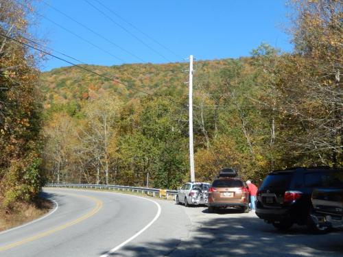 A winding road with parked cars beside a forested area displaying autumn foliage under a clear blue sky.