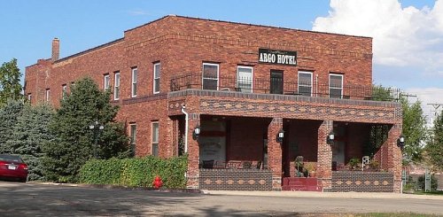 A brick building with a sign reading "Argo Hotel," featuring a porch and surrounded by trees and a clear blue sky.