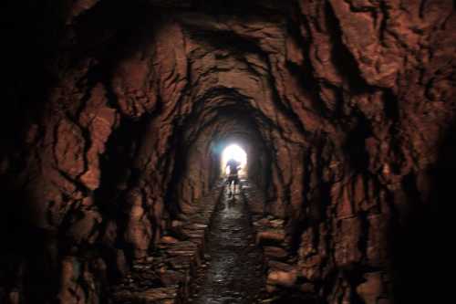 A person walks through a dark cave tunnel, with light visible at the far end. The walls are rugged and rocky.