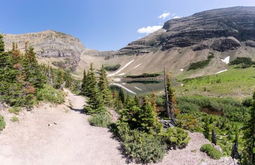 A scenic mountain landscape with a clear lake, surrounded by green trees and rocky hills under a blue sky.