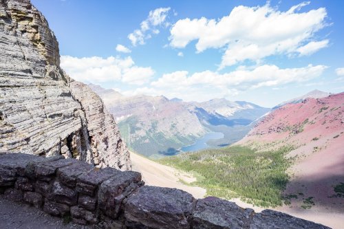 A panoramic view of mountains and a lake under a blue sky with scattered clouds, framed by rocky cliffs in the foreground.