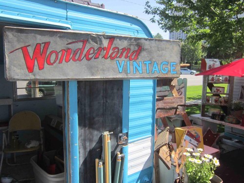 A vintage shop sign reading "Wonderland Vintage" on a blue trailer, surrounded by outdoor decor and greenery.