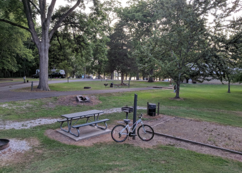 A park scene featuring a picnic table, a bicycle, and green trees in the background, with a gravel path and grassy areas.