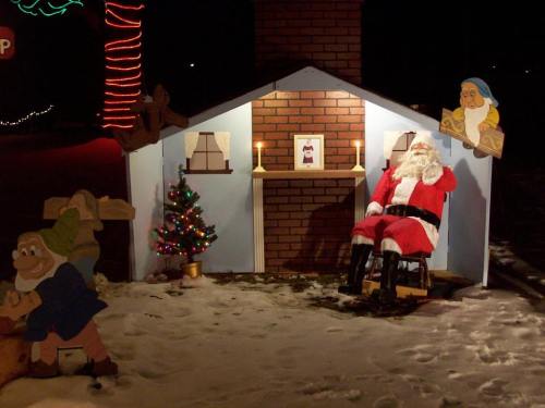 A festive scene with Santa sitting in a chair beside a decorated Christmas tree and a cozy house backdrop.