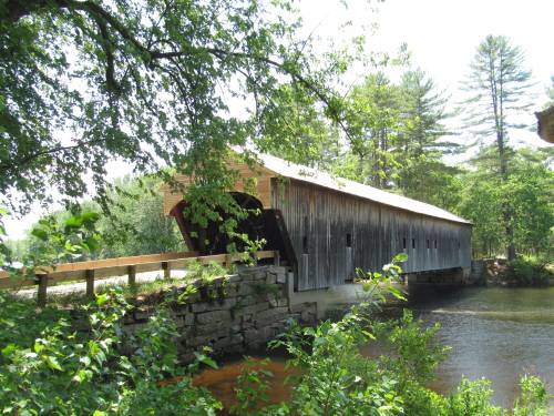 A scenic view of a covered bridge over a river, surrounded by lush greenery and trees on a sunny day.