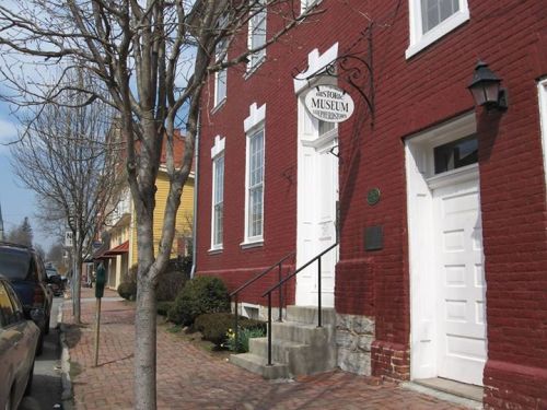A red brick building with a sign reading "History Museum" and a staircase, surrounded by trees and a brick sidewalk.