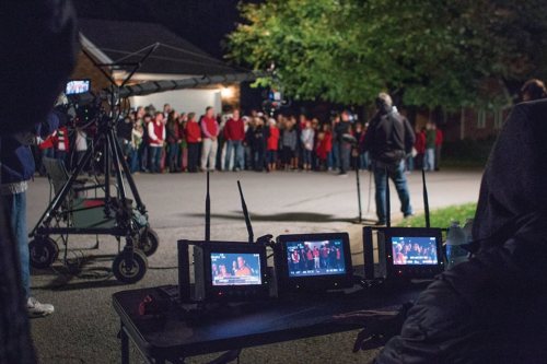 A film crew sets up cameras and monitors in front of a group of people gathered outdoors at night.