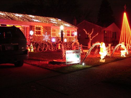 A house decorated with colorful Christmas lights, featuring a large tree, Santa figures, and a sign for a light show.