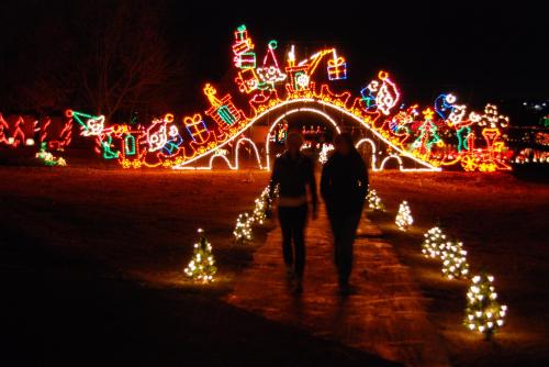 Two people walk along a path lined with small lights, passing under a brightly lit arch decorated for the holidays.
