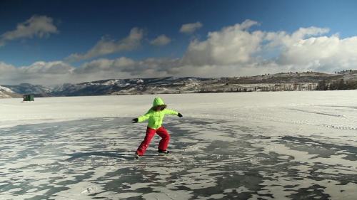 A child in a bright green jacket and red pants ice skating on a frozen lake with mountains in the background.