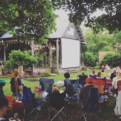 A crowd sits on lawn chairs in a garden, watching a movie on a large outdoor screen surrounded by greenery.