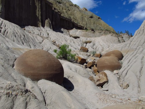 A dry, rocky landscape featuring large, round boulders and textured, sloping terrain under a blue sky.