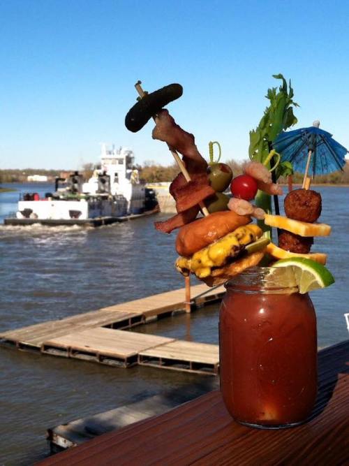 A colorful cocktail garnished with skewered food items, set against a river and a boat in the background.