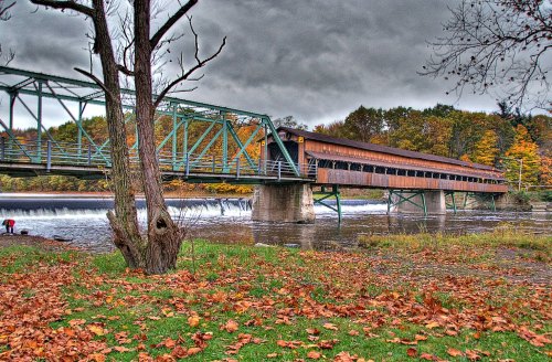 A covered bridge spans a river, surrounded by autumn foliage and fallen leaves under a cloudy sky.