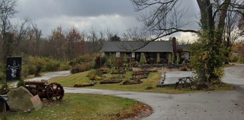 A rustic house with a garden, surrounded by trees, under a cloudy sky. A winding path leads to the entrance.