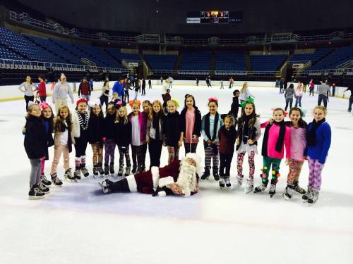 A group of children in skating outfits poses on ice, with one child dressed as Santa lying on the ice.
