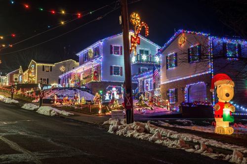 Colorful holiday lights adorn houses along a snowy street, featuring festive decorations and a large inflatable Santa Snoopy.