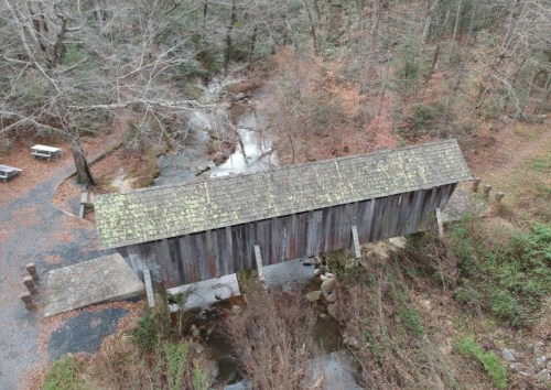 Aerial view of a rustic covered bridge over a small stream, surrounded by bare trees and autumn foliage.