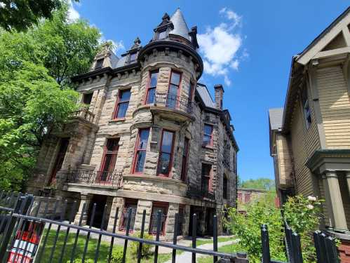 A historic stone mansion with a turret, red accents, and a lush green yard under a bright blue sky.