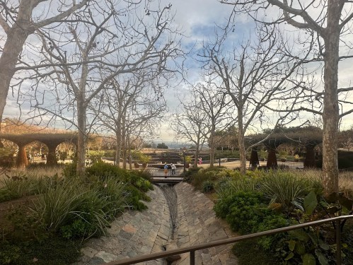 A serene park scene with bare trees, a stone pathway, and people walking in the distance under a cloudy sky.