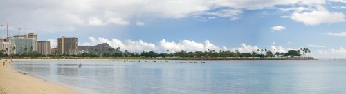 Panoramic view of a beach with calm water, palm trees, and city skyline under a partly cloudy sky.