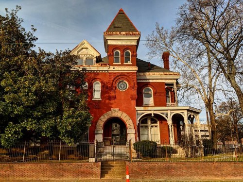 A large, red brick Victorian house with a tower, surrounded by trees and a wrought-iron fence, under a clear sky.