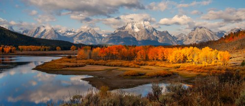 A serene landscape featuring a lake, vibrant autumn trees, and majestic mountains under a partly cloudy sky.