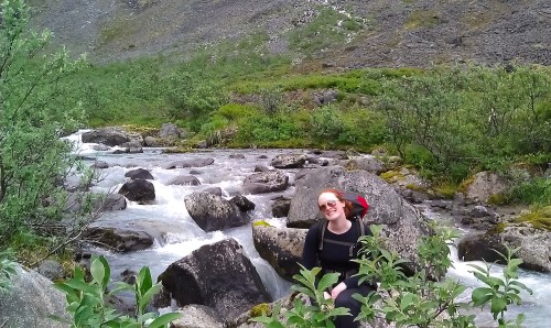A person sits by a flowing river surrounded by rocks and greenery in a mountainous landscape.