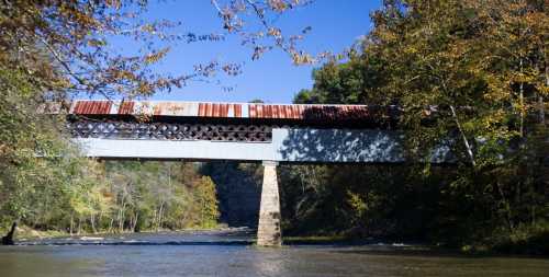 A rusted train bridge spans a river, surrounded by trees with autumn foliage under a clear blue sky.