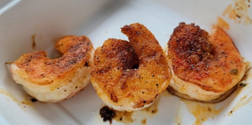 Three seasoned, grilled shrimp on a white takeout container, showcasing a golden-brown crust.