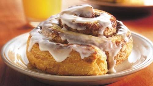 A freshly baked cinnamon roll topped with creamy icing on a plate, with a glass of orange juice in the background.
